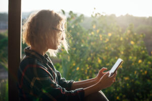 Uma garota loira usa um smartphone no telhado de um prédio contra o fundo de árvores à luz do sol. O conceito de trabalho e entretenimento fora do escritório, ao ar livre durante a viagem.